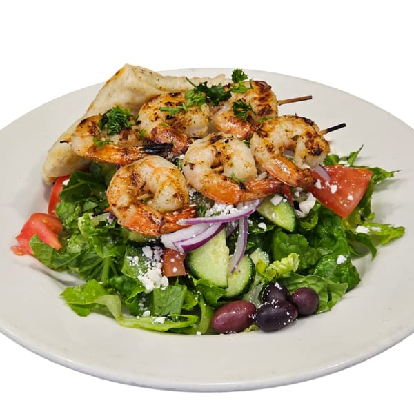 Grilled shrimp skewers on a Greek salad with feta, olives, tomatoes, and pita bread.
