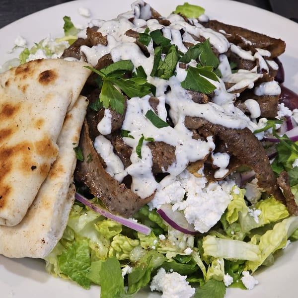 Greek gyro meat with tzatziki sauce, feta, lettuce, red onion, and pita bread on a white plate.