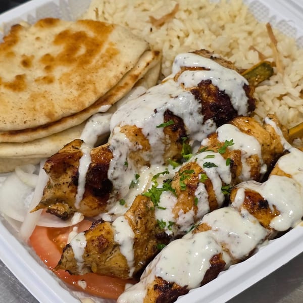 Greek chicken souvlaki skewers with tzatziki, rice, pita bread, tomatoes, and onions in a white container.