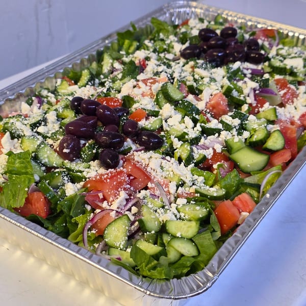 A large catering tray of Greek salad with lettuce, tomatoes, cucumbers, feta, red onion, and olives.